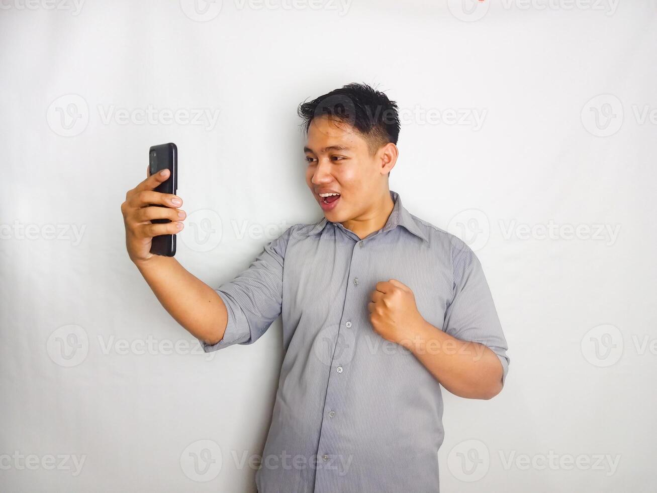 Young Asian man showing wow face expression when looking to his mobile phone wearing grey shirt isolated on white background photo