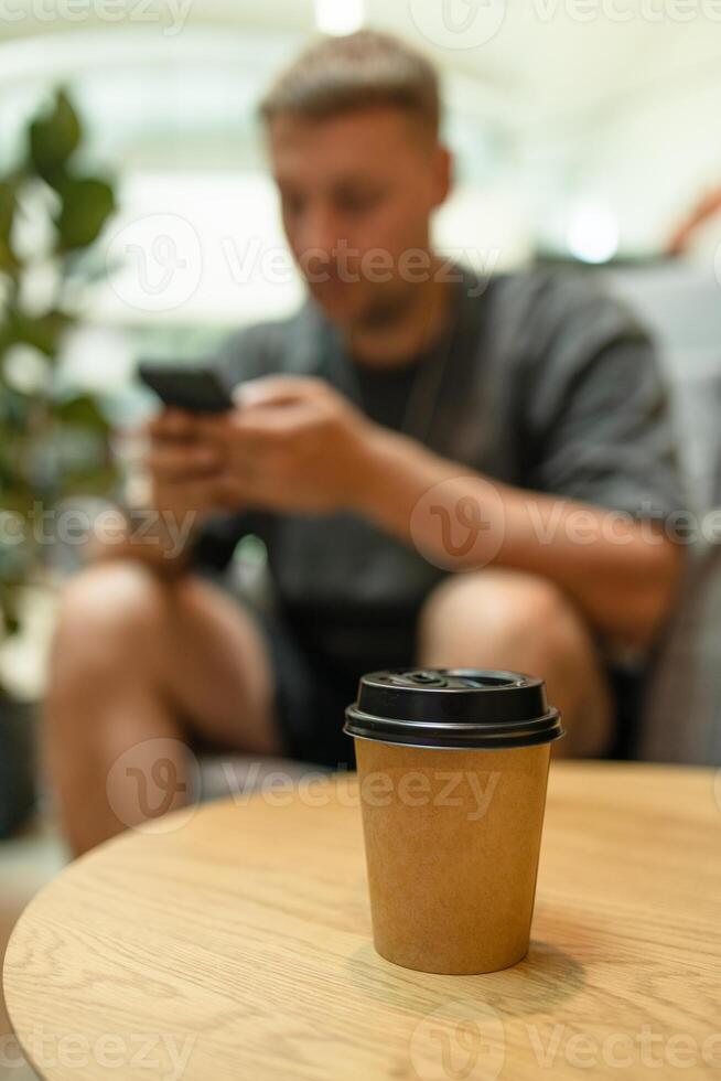 Takeaway paper coffee cup with aroma latte coffee on a wood table at city cafe. Male with social media addiction on smartphone scrolling internet, enjoy virtual life rest look at screen. photo