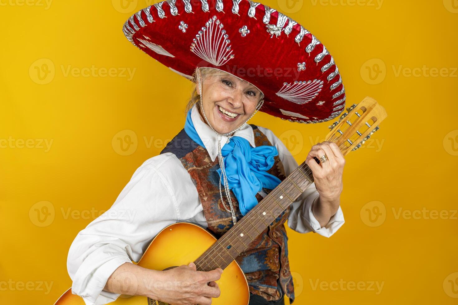 portrait of a happy middle aged woman smiling in front of a yellow background enjoying playing guitar dressed as a Mexican mariachi with sombrero photo