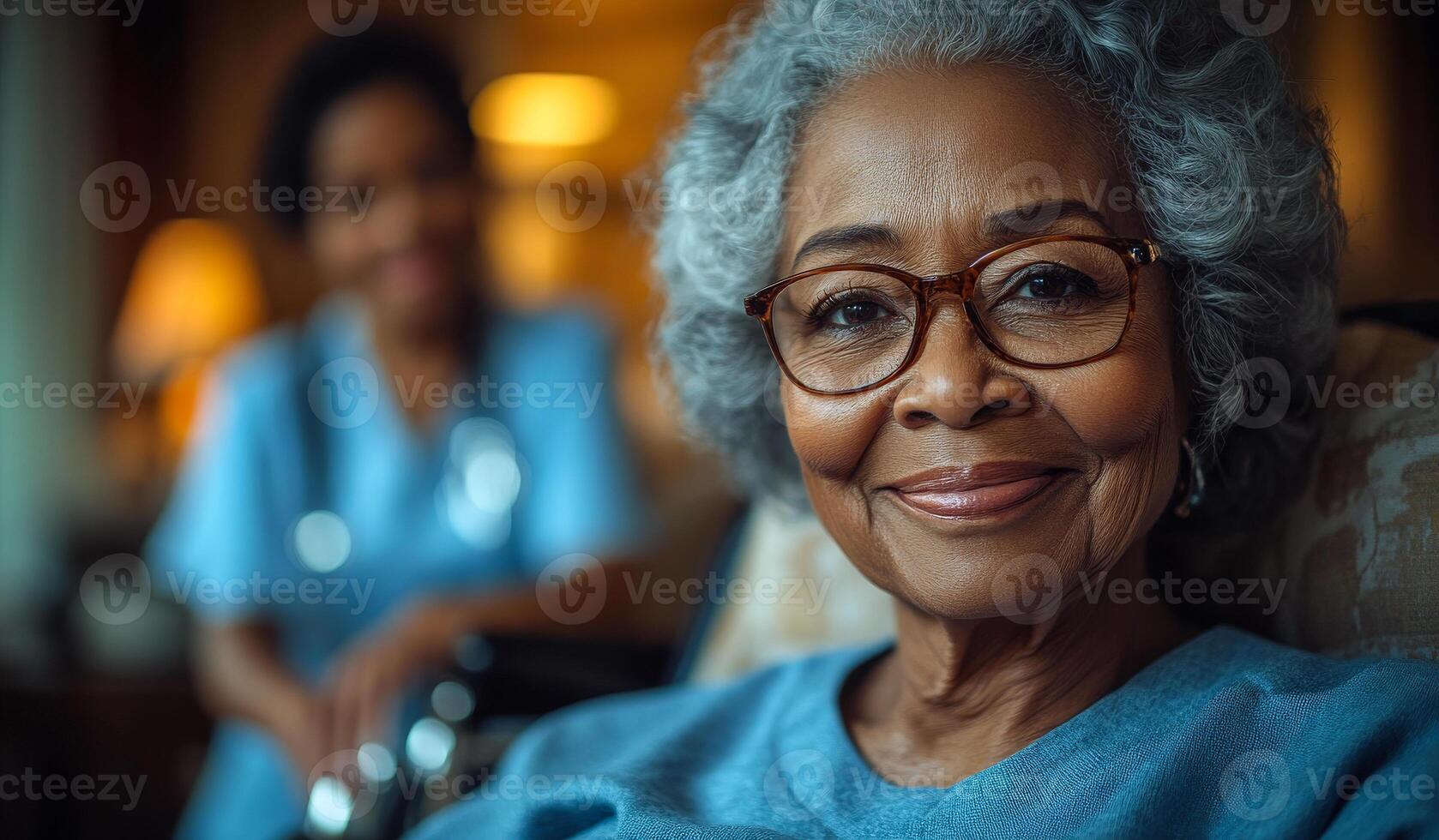 A woman in a blue shirt and glasses is smiling. She is sitting in a chair. A nurse is standing behind her photo