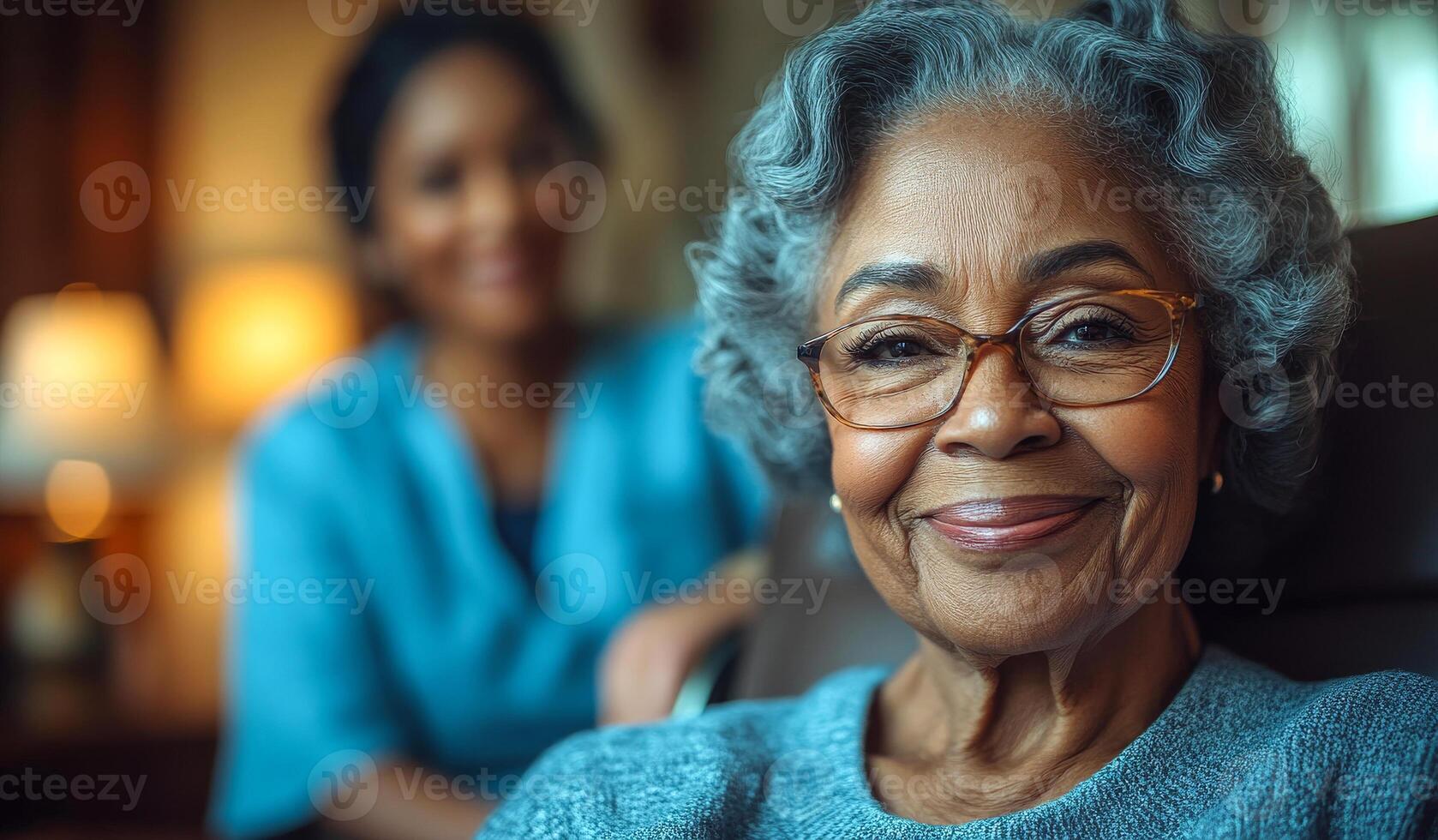 A woman with glasses is smiling and sitting in a chair. A woman in a blue shirt is standing behind her photo