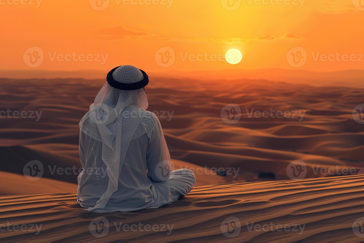 Man wearing traditional clothing sitting on sand dune watching sunset over desert photo