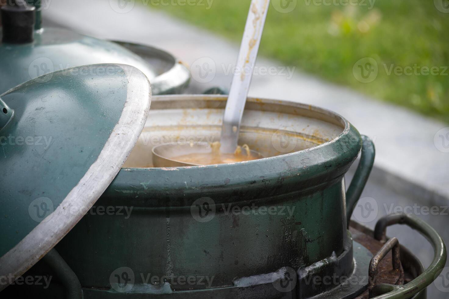 Large green metal pot with open lid and ladle serving soup outdoors on a rustic cooking setup photo