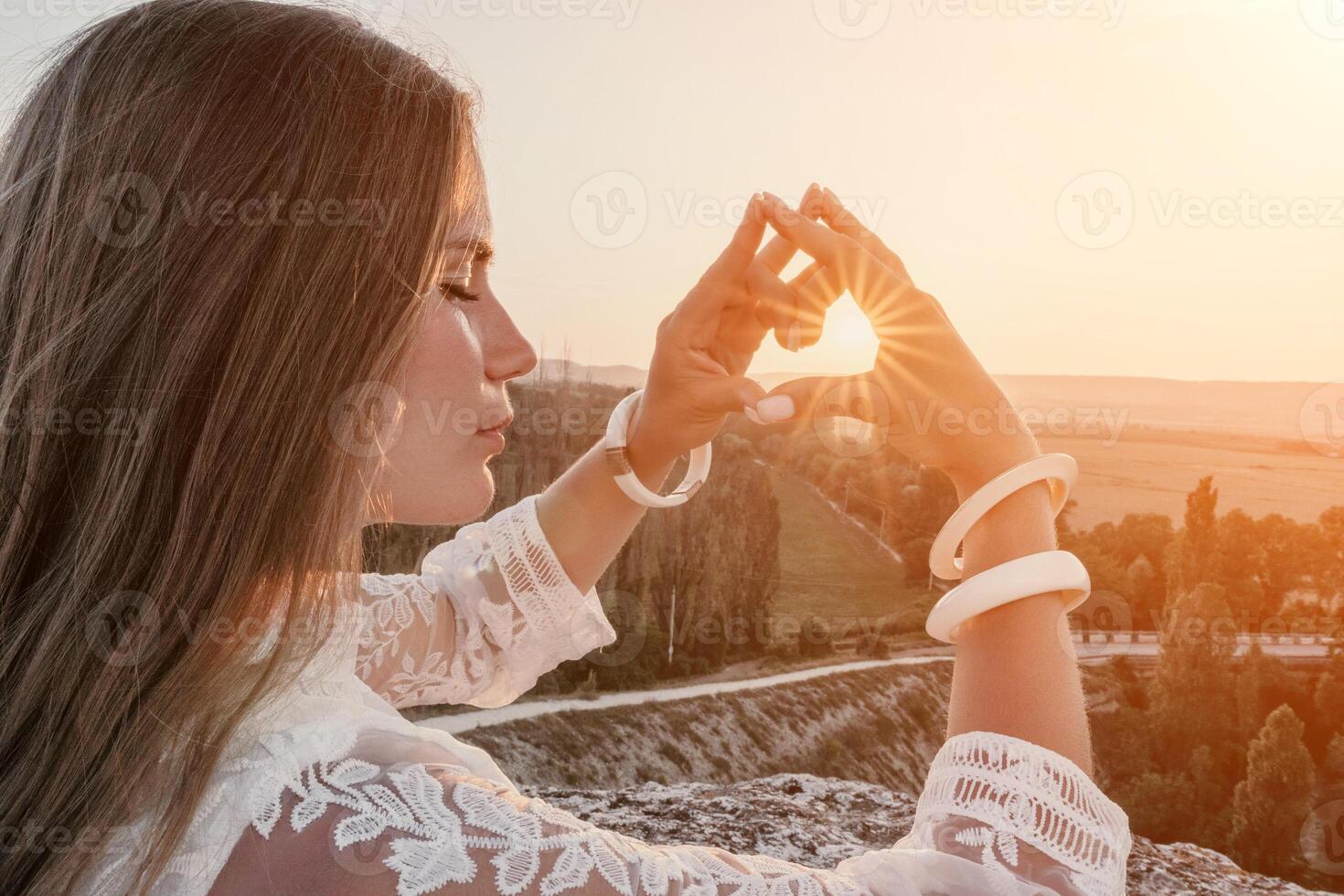 Happy woman in white boho dress making heart sign with hands on sunset in mountains. Romantic woman with long hair standing with her back on the sunset in nature in summer with open hands. photo