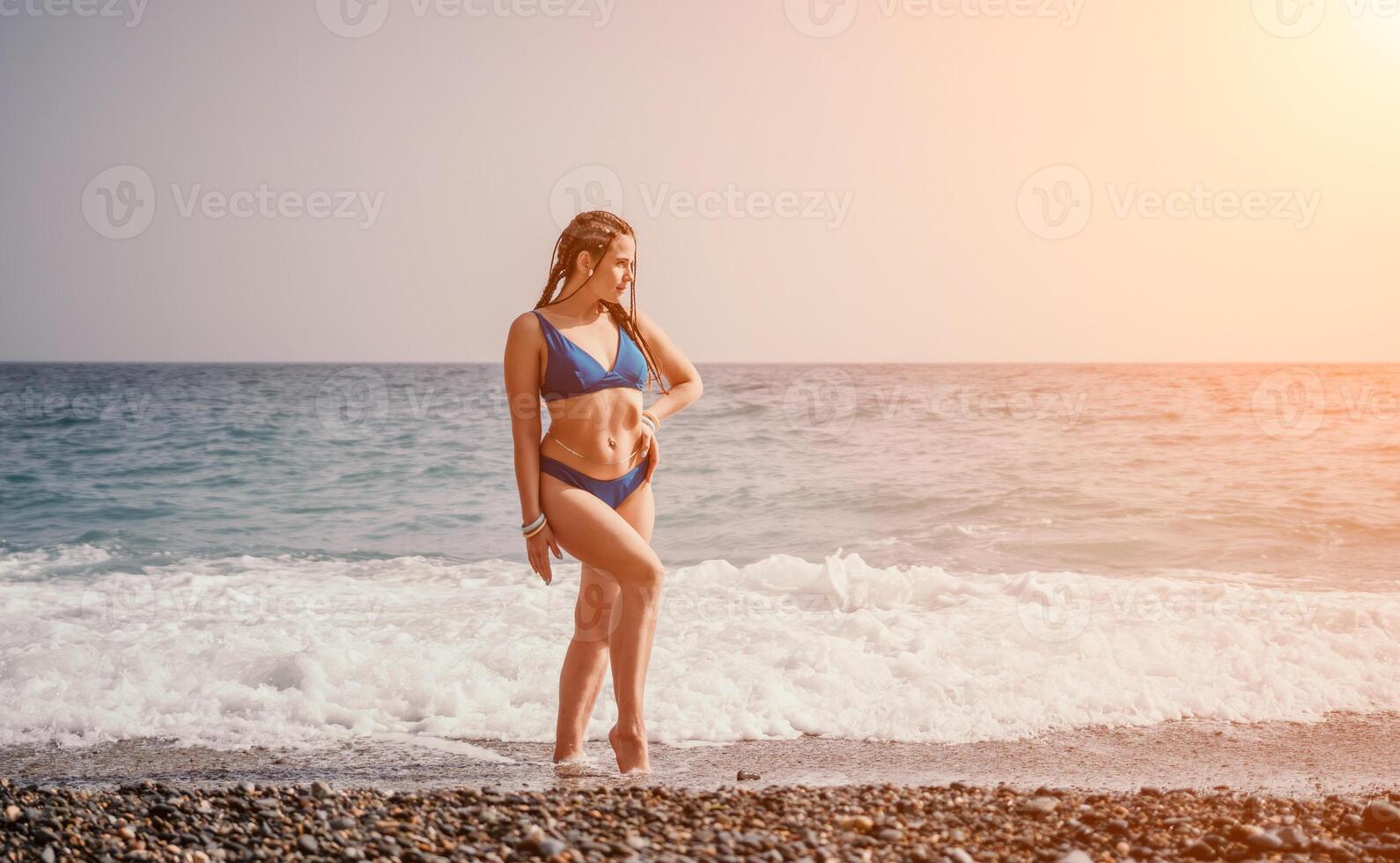 A tourist woman in blue bikini on beach enjoys the turquoise sea during her summer holiday. Rear view. Beach vacation. woman with braids dreadlocks standing with her arms raised enjoying beach ocean. photo