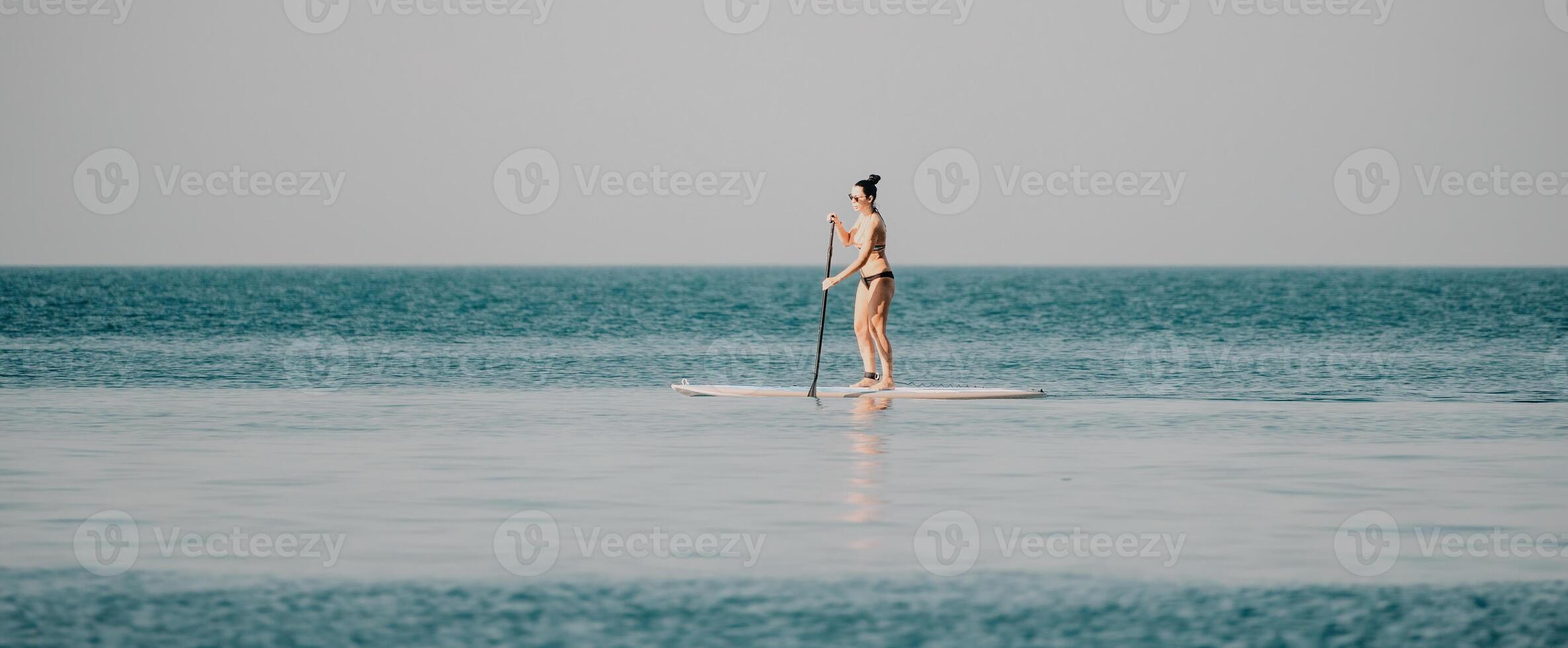 Sea woman sup. Silhouette of happy middle aged woman in rainbow bikini, surfing on SUP board, confident paddling through water surface. Idyllic sunset. Active lifestyle at sea or river. photo