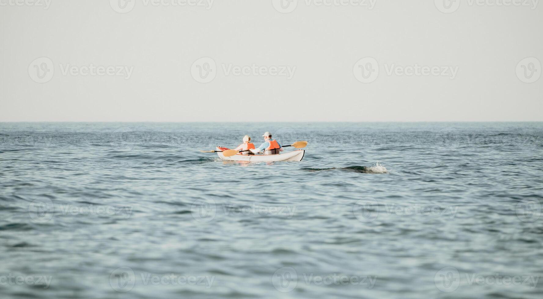 Happy couple kayaks in an inflatable kayak on the sea at sunset. Couple kanoeing in the sea near the island with mountains. People kayaking in life jackets sail. Back view photo