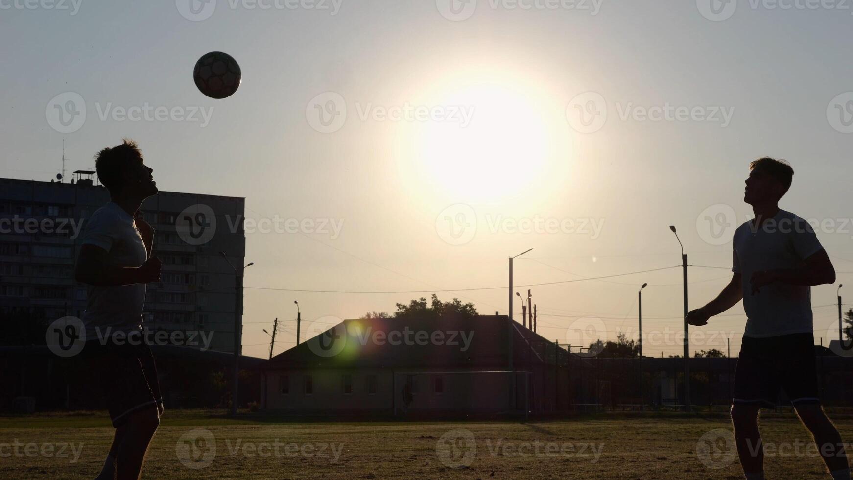 Silhouette of professional footballers kicking soccer ball with their heads on stadium at sunset. Two sportsmen showing tricks with ball while passing it to each other at field. Freestyle football photo