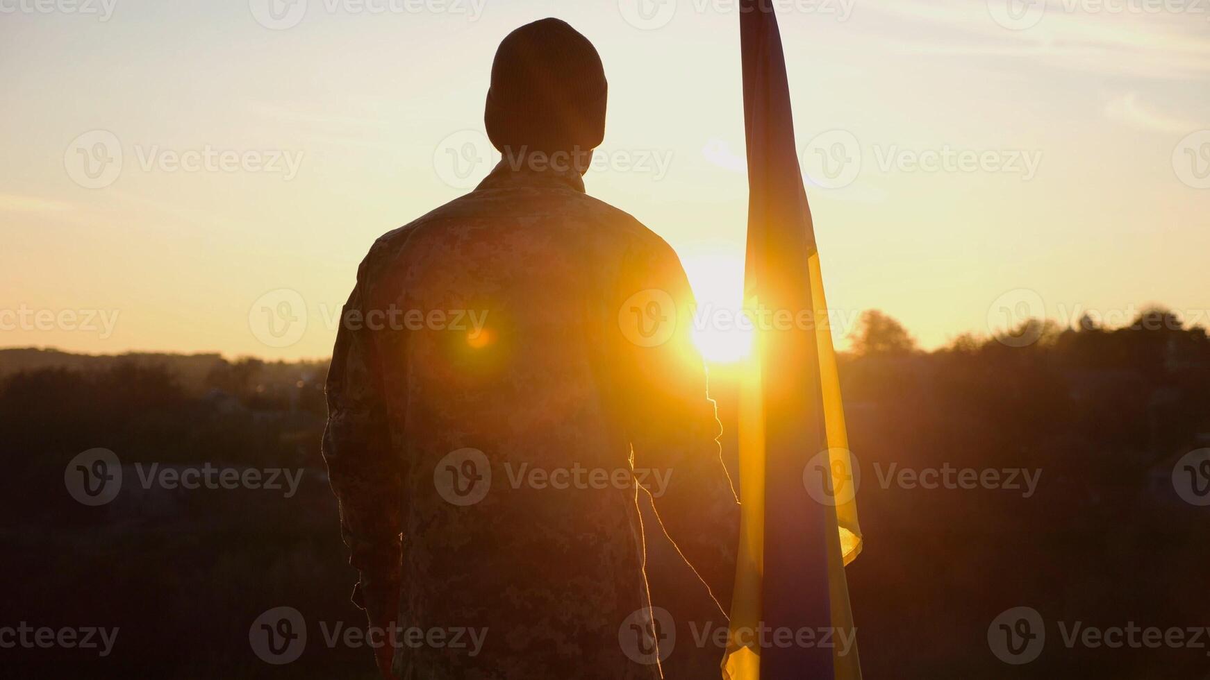 Male ukrainian army soldier stands with national banner against background of beautiful sunset. Young man in camouflage uniform with Ukraine flag as symbol of victory against russian aggression photo