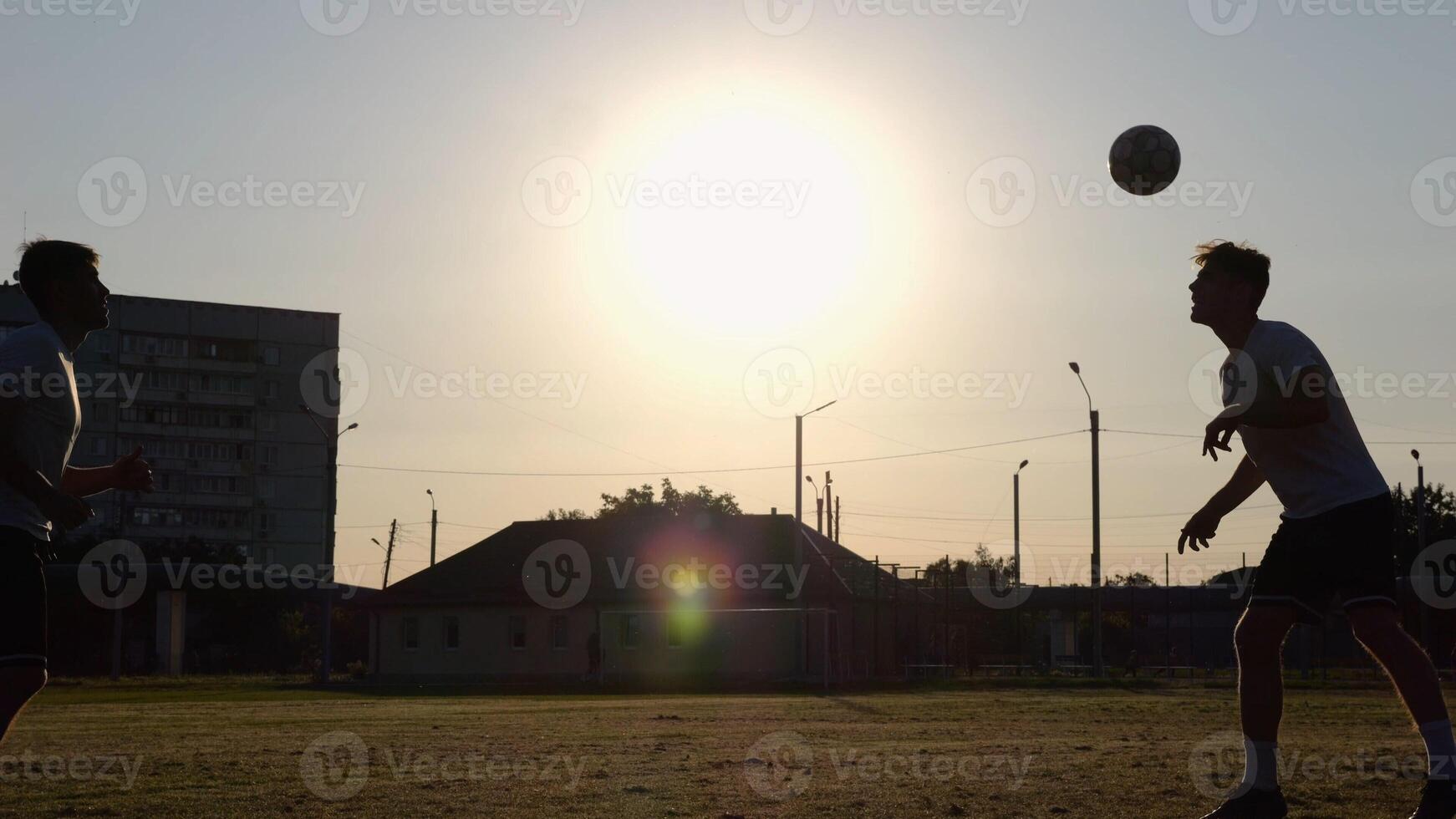 Silhouette of professional footballers kicking soccer ball with their heads on stadium at sunset. Two sportsmen showing tricks with ball while passing it to each other at field. Freestyle football photo