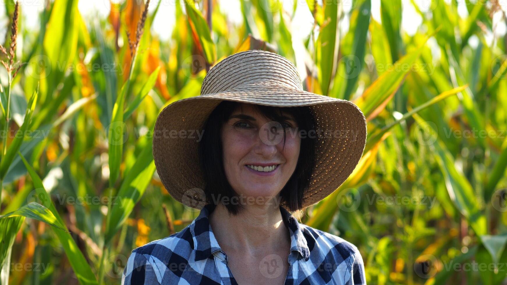 Happy smiling female farmer looks into camera standing near corn field. Portrait of adult beautiful agronomist in straw hat with maize meadow at background. Agricultural business concept. Close up photo