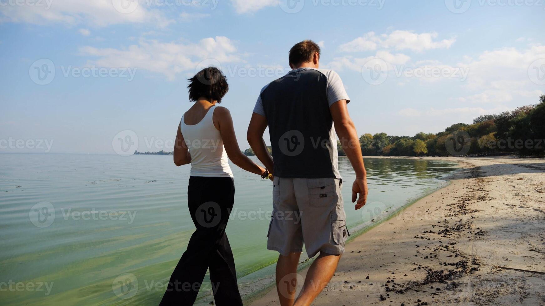 Couple holding hands of each other and walking at coast. Happy pair going along seashore at sunny day. Lovers spending time together at resort. Concept of vacation or holiday. Slow mo photo