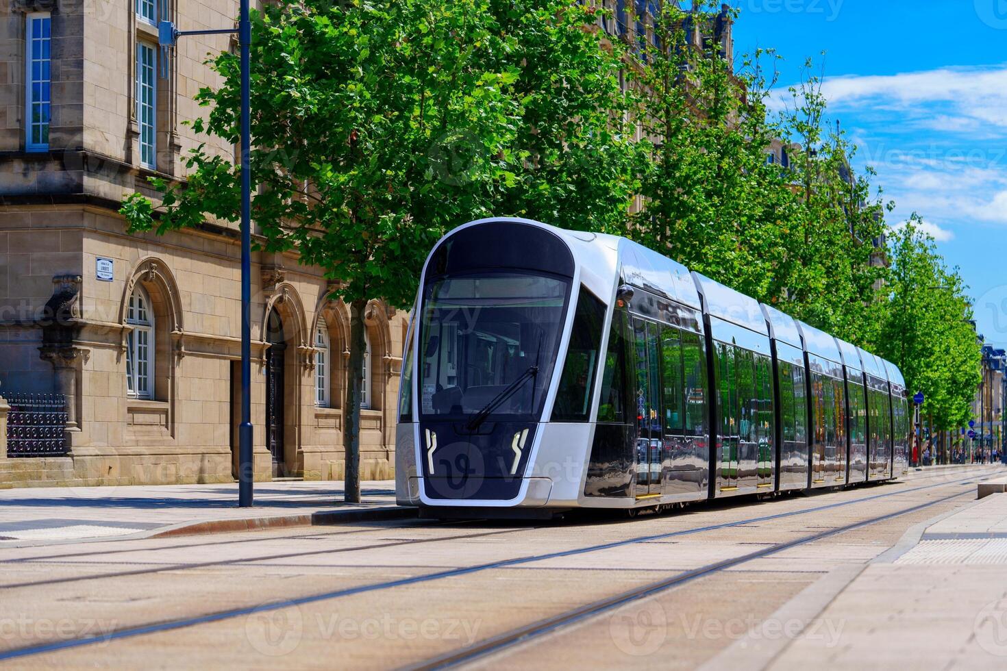 Luxembourg, A modern silver tram moves swiftly along the street, creating a motion blur, while a vintage stone building with many windows stands alongside, surrounded by trees and urban scenery. photo