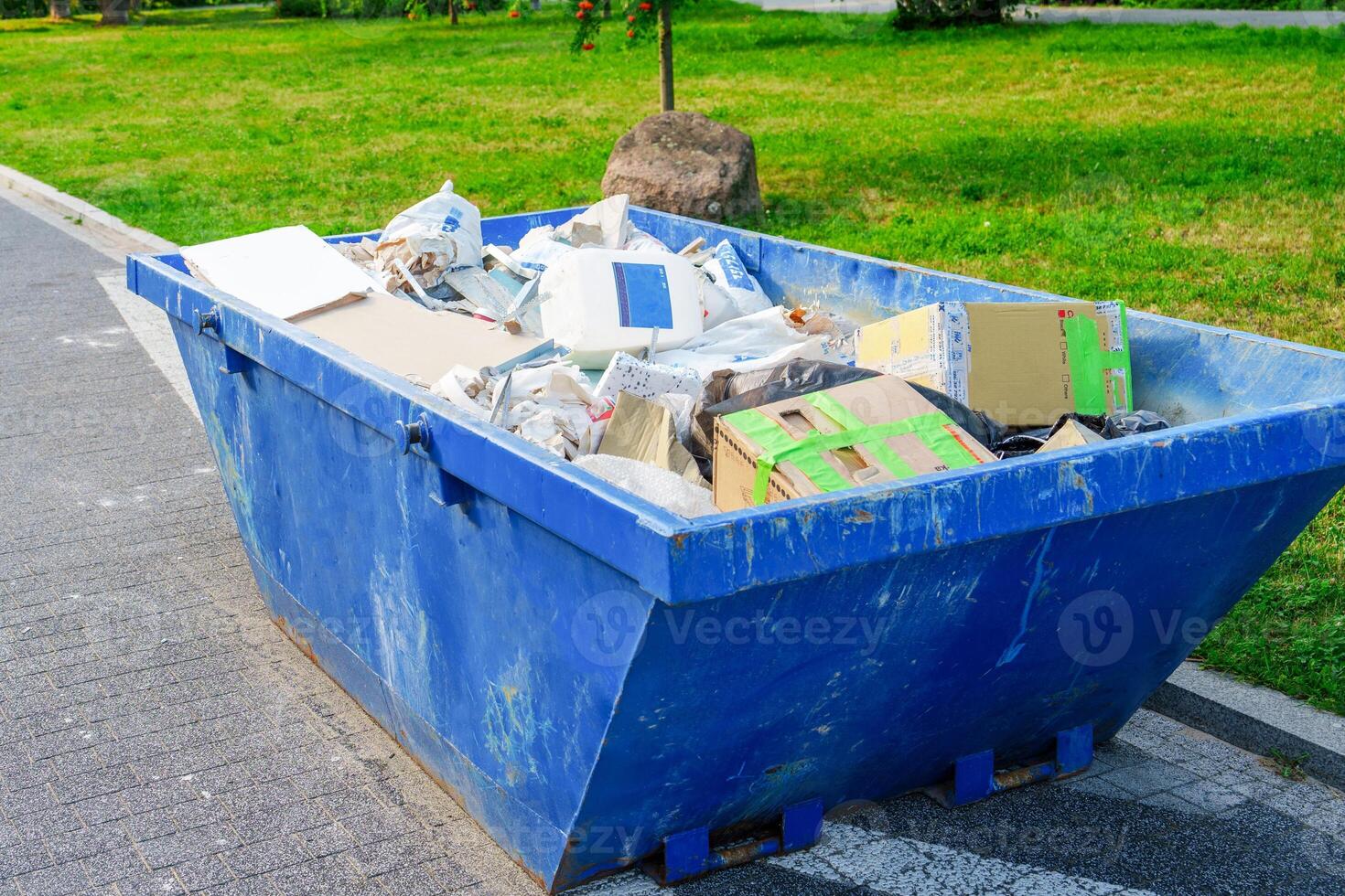 A blue dumpster is sitting in the park. The dumpster is filled with various types of garbage including cardboard boxes, paper, and plastic bottles. The dumpster is sitting on a paved path next to the grass photo