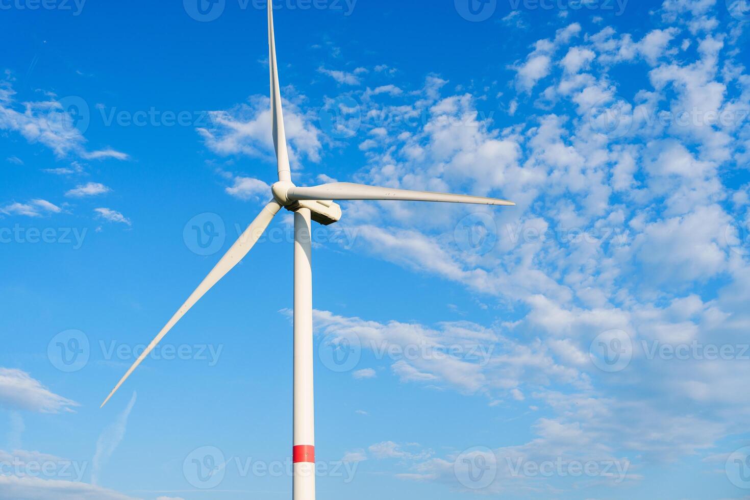 A lone wind turbine stands tall against a blue sky with fluffy clouds. The turbines white blades spin slowly, capturing wind energy and converting it into clean electricity. photo