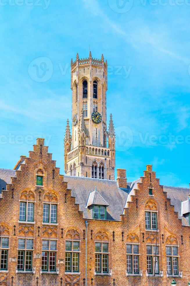 Bruges, Belgium, The clock tower is an architectural marvel in the town square, made of red brick with detailed carvings. Its large clock face gleams under the clear blue sky, creating a charming and picturesque scene. photo