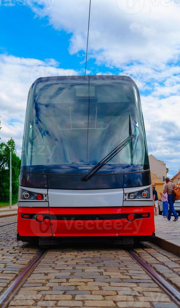 The tram is sleek and modern, featuring a large windshield and a vibrant red body. It glides along a brick track, surrounded by blurred buildings, people walking by, and a lively urban atmosphere. photo