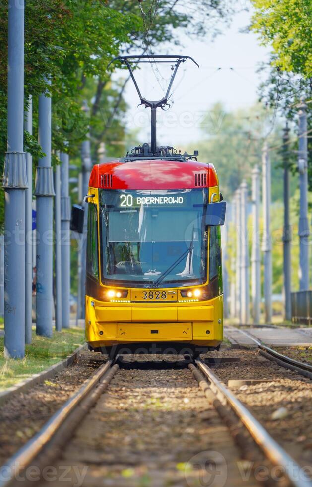 The image features a modern yellow and red tram on a bustling streetof Warsaw. The tram is sharply in focus against a blurred background, emphasizing its vibrant colors and creating a dynamic sense of motion. photo