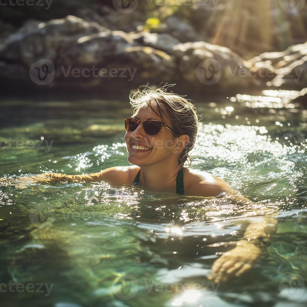 Smiling woman swimming in clear water on a sunny day, wearing sunglasses and enjoying nature in a serene environment photo