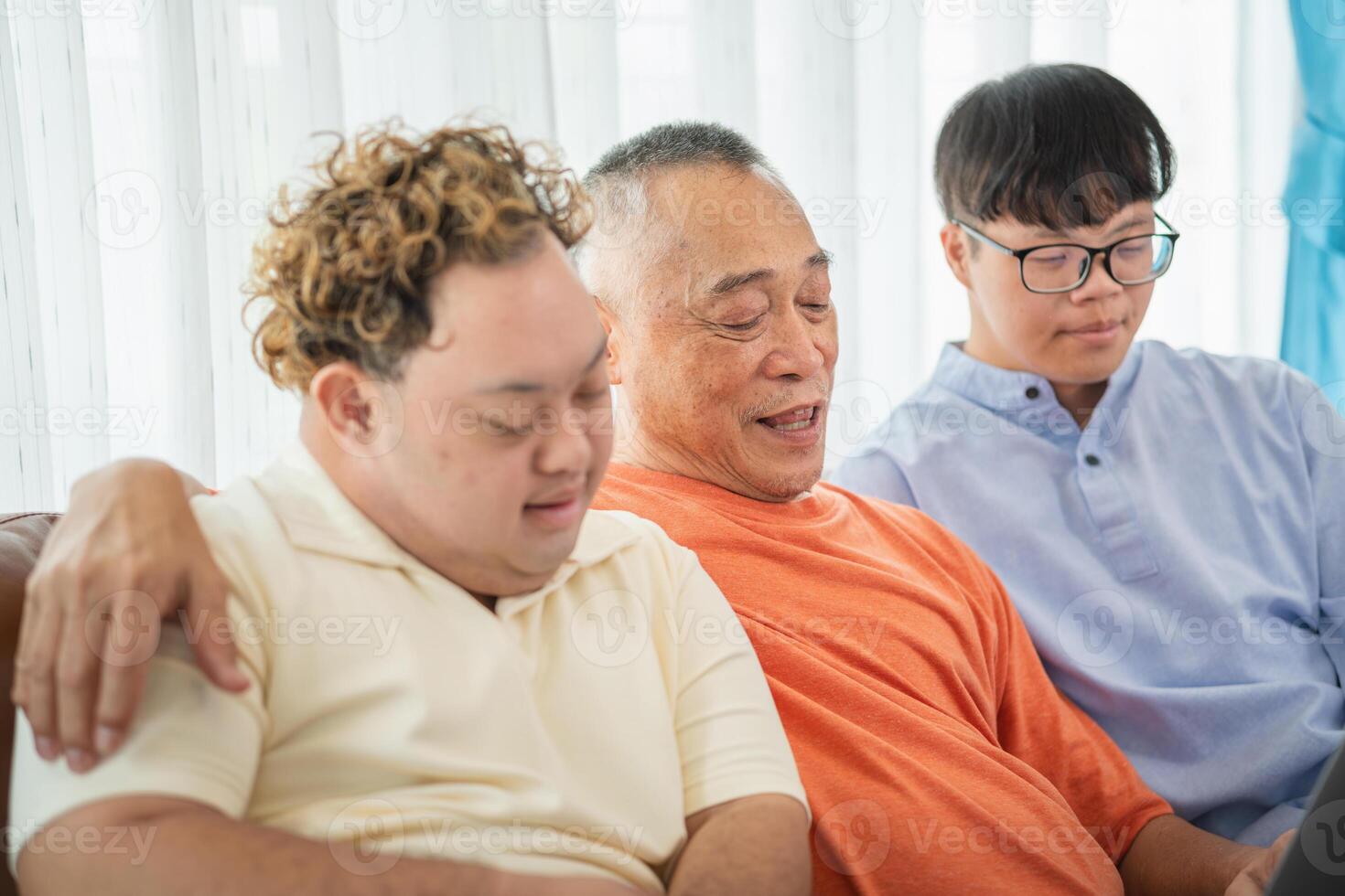 Senior man having a good time sitting on couch at home with adult son, Asian grandfather and grandson using laptop doing a call in living room photo