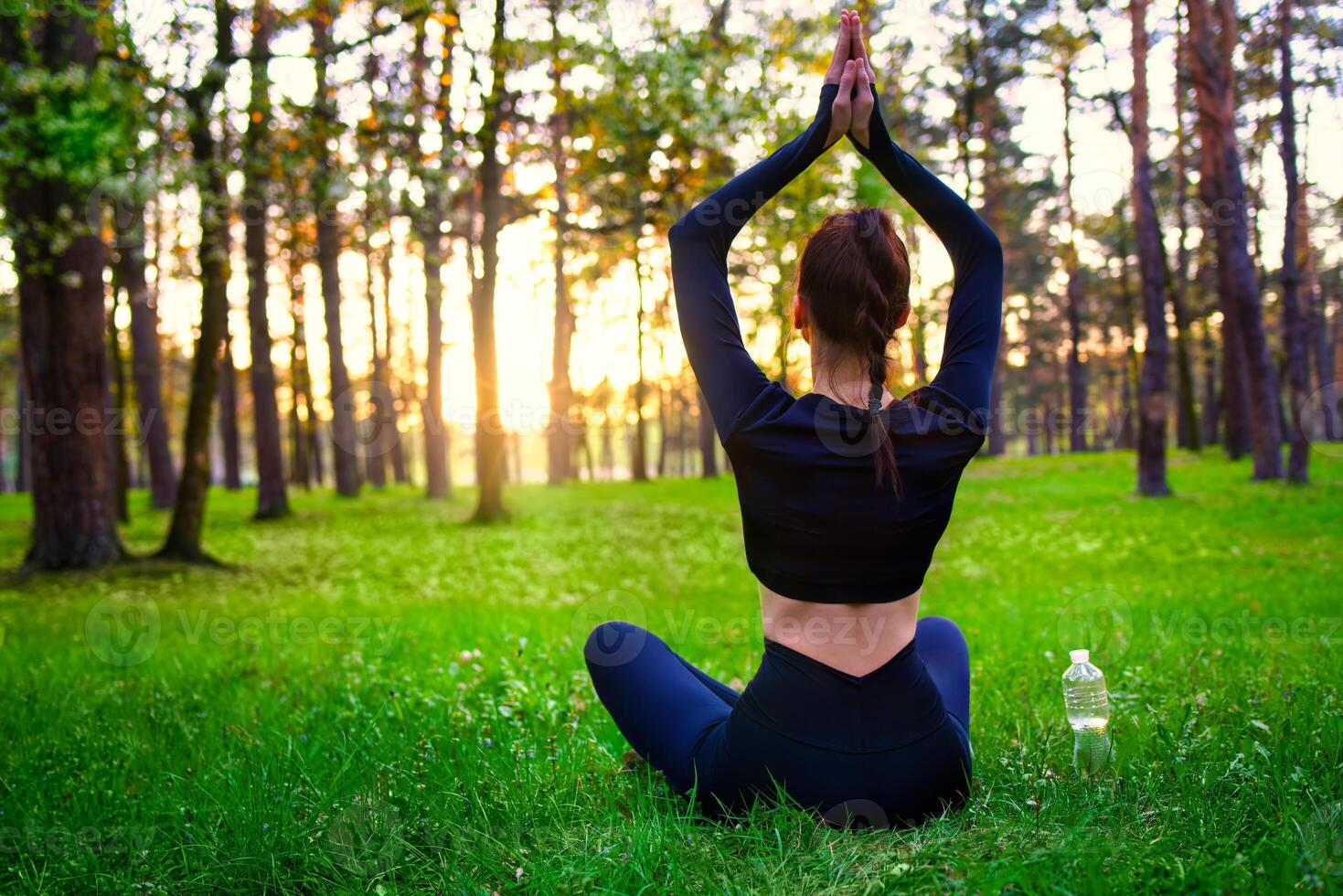 Slim beautiful woman in black tight fitting tracksuit sitting on grass in forest in yoga pose. back view photo