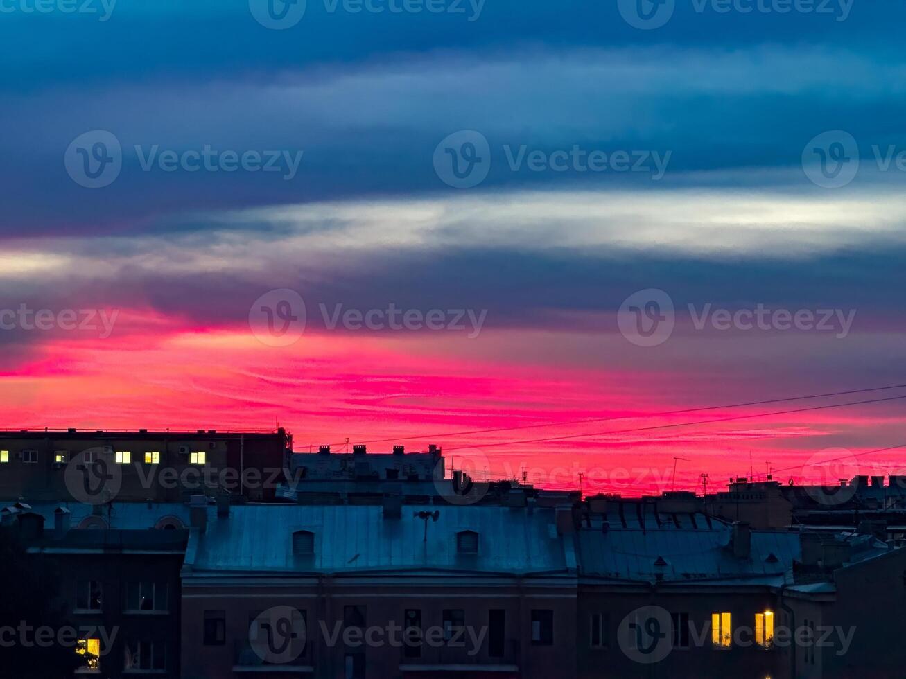 A stunning sunset scene with deep pink and blue hues in the sky, silhouetting city rooftops. A few lit windows in the darkening buildings add warmth and contrast to the vibrant evening sky photo