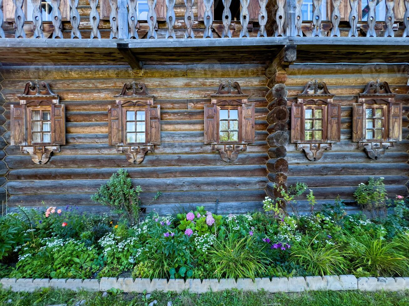 A close-up of beautifully carved wooden windows on a traditional Russian log house , izba. The ornate shutters, detailed woodwork, and charming flower bed create a picturesque, rustic ambiance photo