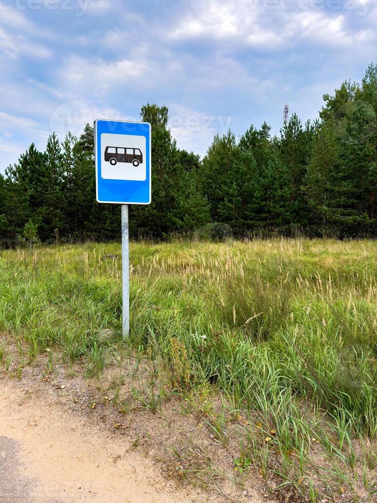 A bus stop sign stands on the side of a quiet rural road, surrounded by grassy fields and a backdrop of dense pine trees. The blue sky with scattered clouds adds a peaceful atmosphere to the scene photo