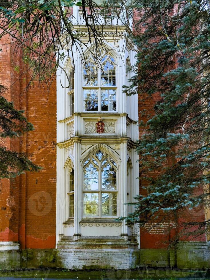 A close up of a beautifully preserved Gothic window from a historic estate, framed by red brick and lush pine branches. The intricate architectural details and glass reflections emphasize the building photo