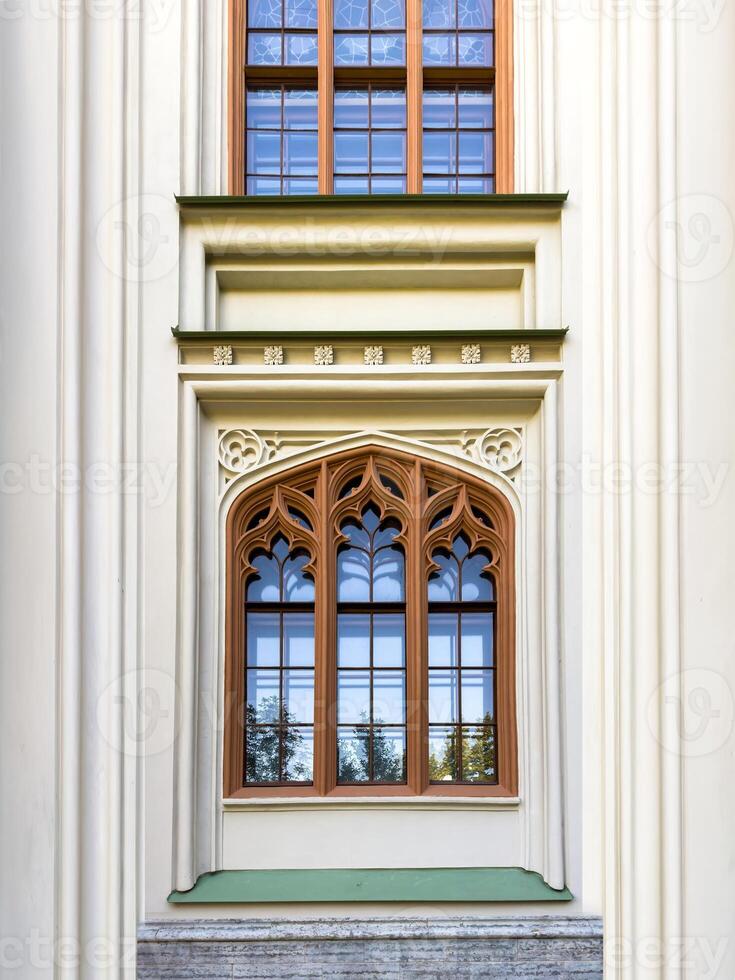 A close up of a beautifully preserved Gothic window from a historic estate, framed by red brick and lush pine branches. The intricate architectural details and glass reflections emphasize the building photo