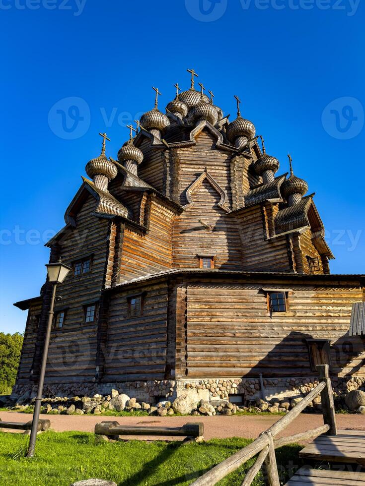 A traditional wooden Orthodox church with intricate onion domes, basking in the warm sunlight on a clear day. The rustic architecture stands against a vibrant blue sky, highlighting the cultural and photo