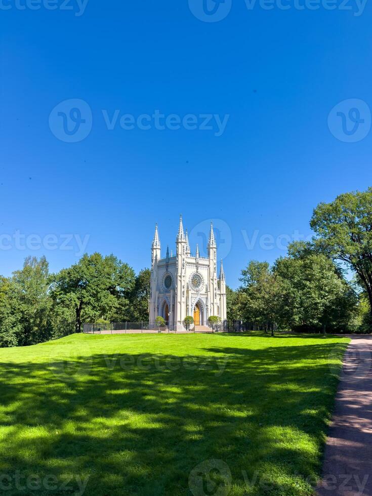 A stunning Gothic-style chapel standing in the middle of a green park under a bright blue sky. Captured from a wide-angle and close-up perspective, the chapel's intricate details and pointed spires photo