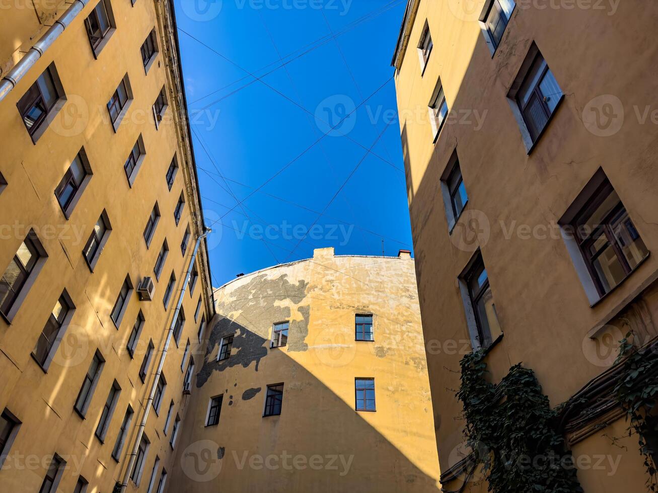 A photo of a secluded courtyard surrounded by tall, yellow apartment buildings. Sunlight casts shadows on the walls, highlighting the weathered textures of the facade and creating a peaceful urban