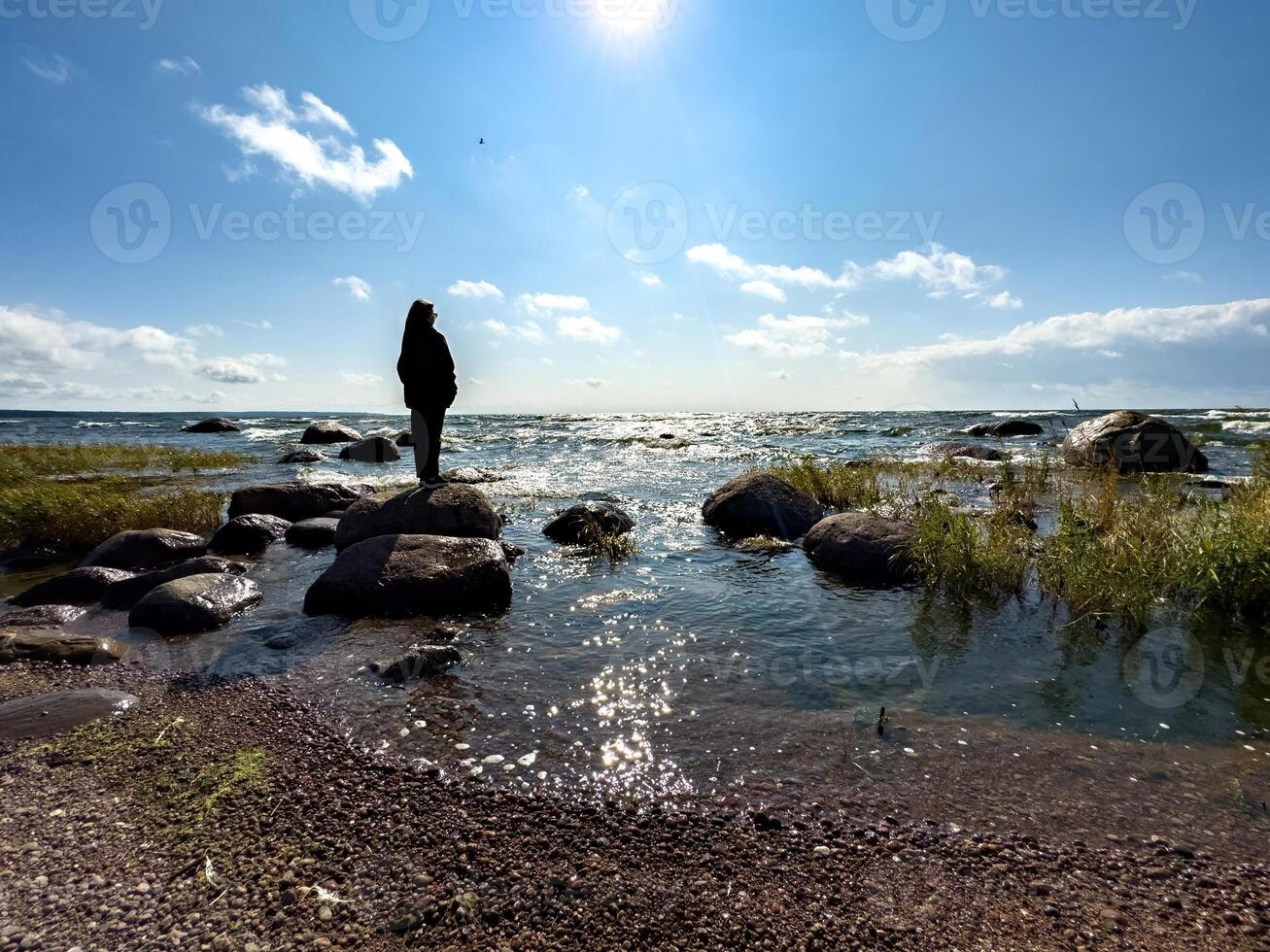 A woman wearing a hoodie stands on large rocks by the shore, gazing at the sea under the bright sunlight. Waves gently crash against the rocks while the sun casts sparkling reflections on the water photo