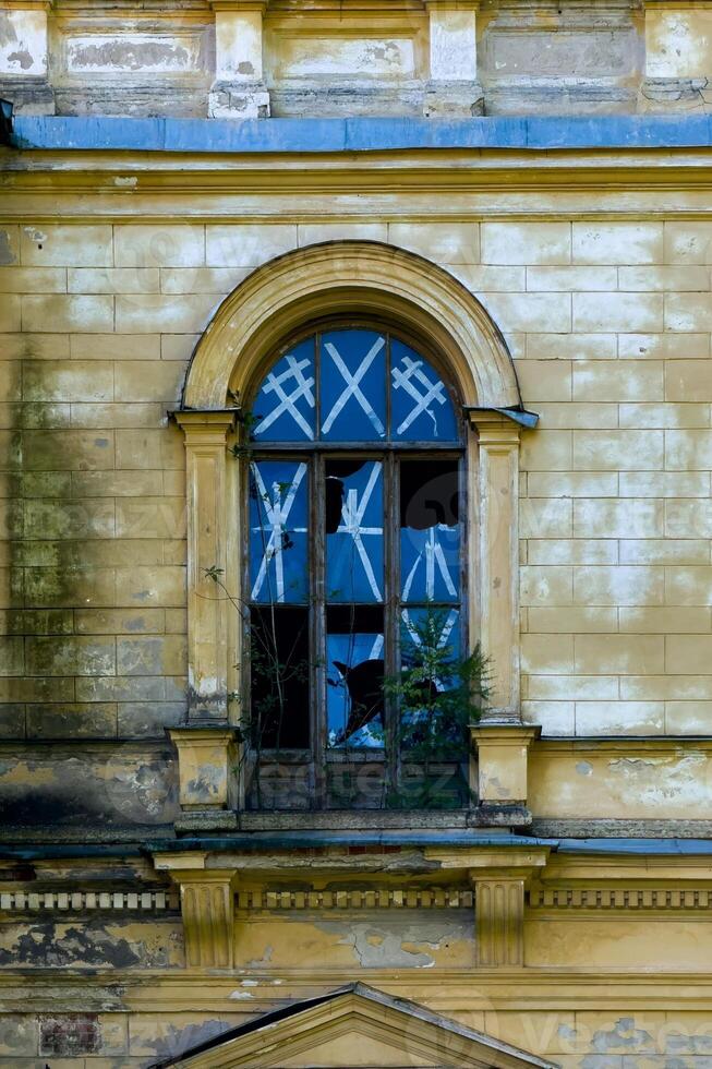 A haunting image of a broken arched window in an abandoned mansion. The peeling yellow paint of the building contrasts with the shattered glass panes and crisscrossing tape. Overgrown plants peek photo