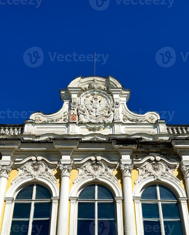 The facade of an abandoned Baroque estate in Peterhof, Russia, with its once-glorious architecture now showing signs of decay. A wide-angle shot reveals intricate, weathered details under a bright photo