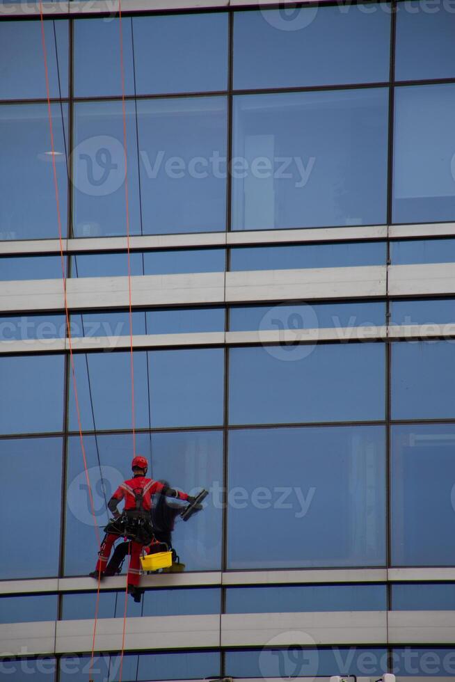 Industrial climber at difficult job, vertical photo of climber washing windows of glass skyscraper, cleaning exterior of modern building