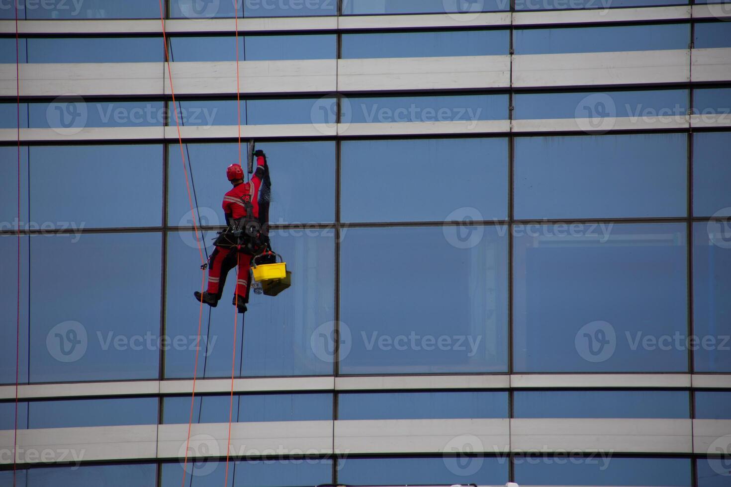 Industrial climber in red overalls cleans the glass windows of skyscraper with office space, hard work at height for cleanliness of building exterior photo