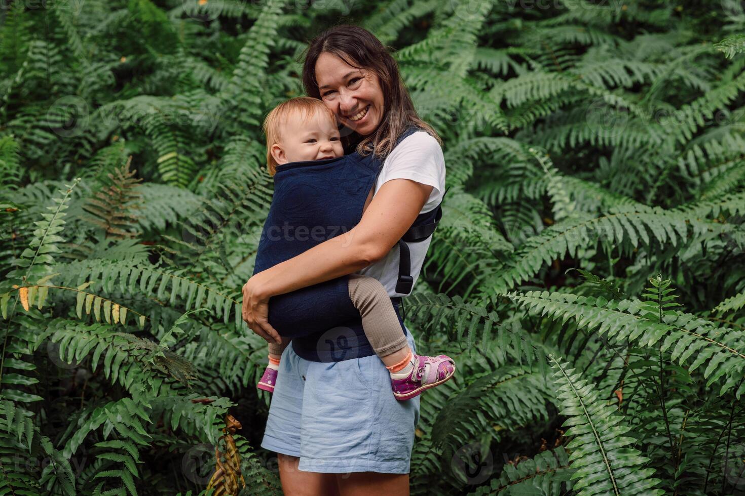 Cheerful mother with her cute baby daughter in park between fern leaves photo