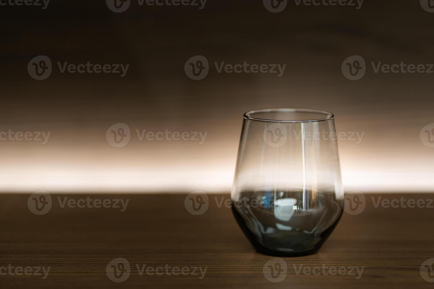 A single, empty glass cup sits on a dark wooden table. The glass has a curved bottom and a wide rim. The background is a gradient of dark brown to a lighter brown. photo