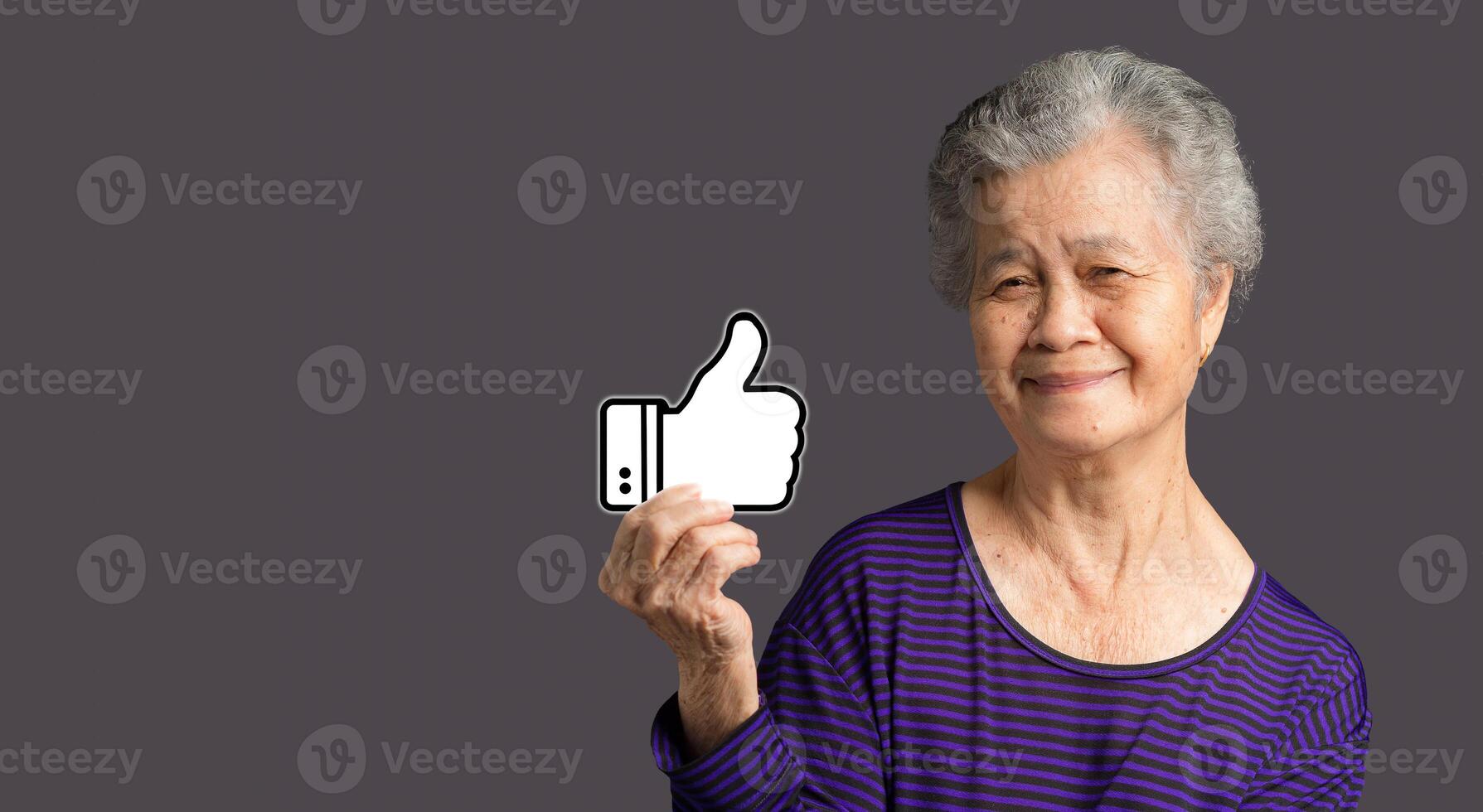 Service rating, satisfaction concept. Portrait of an elderly Asian woman holding a thumbs up a symbol and looking at the camera with a smile while standing on a gray background photo