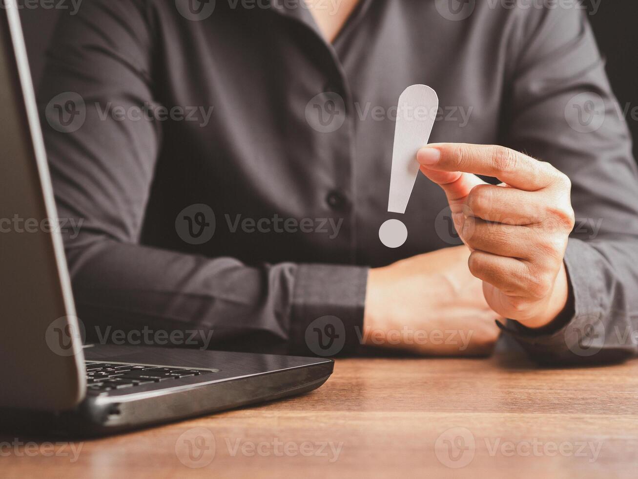 Exclamation mark concept. Hand of a businessman in a black shirt holding a white exclamation mark while sitting in the office. photo