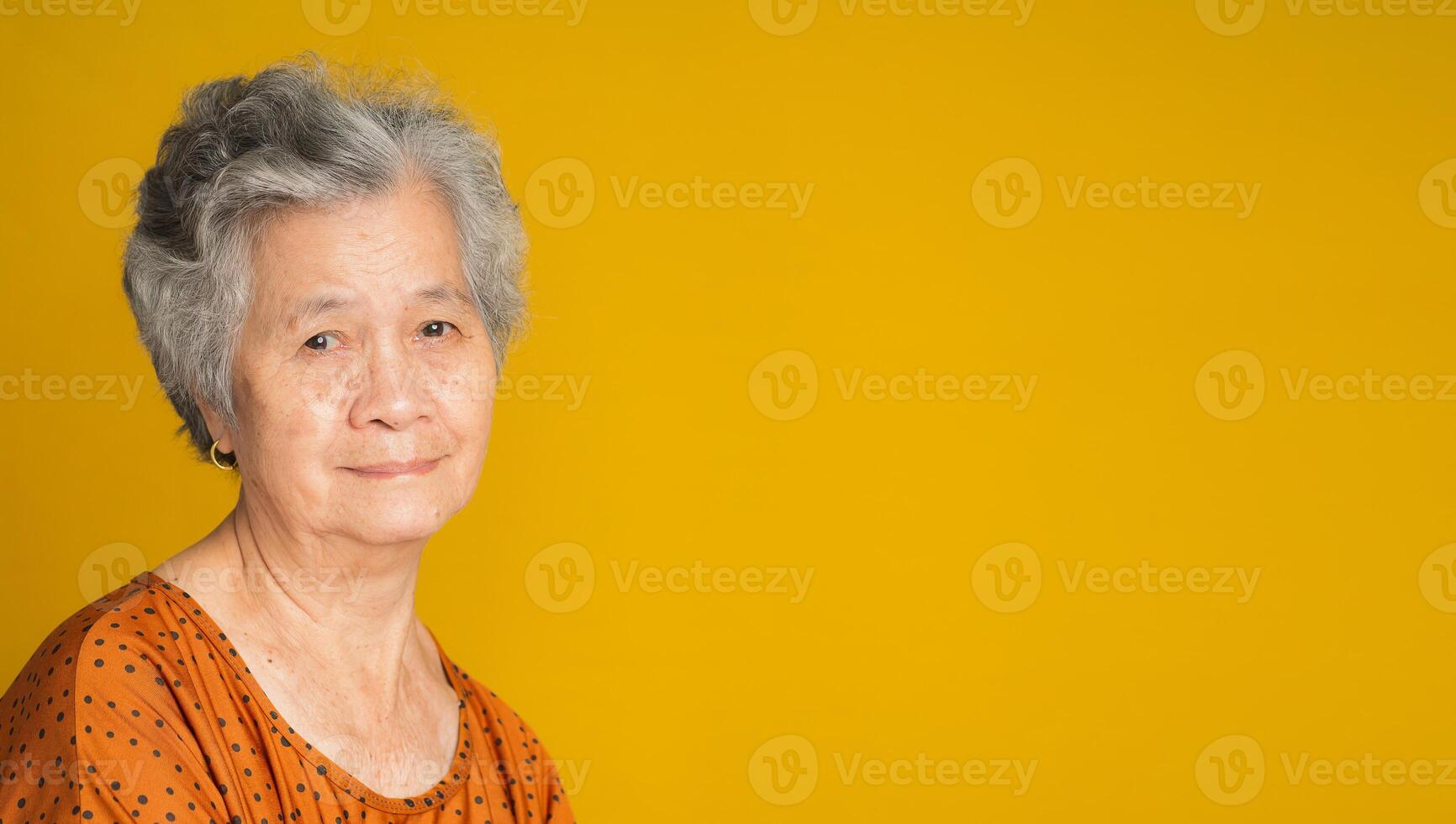 An elderly Asian woman with short gray hair looking at the camera with a smile while standing on a yellow background photo