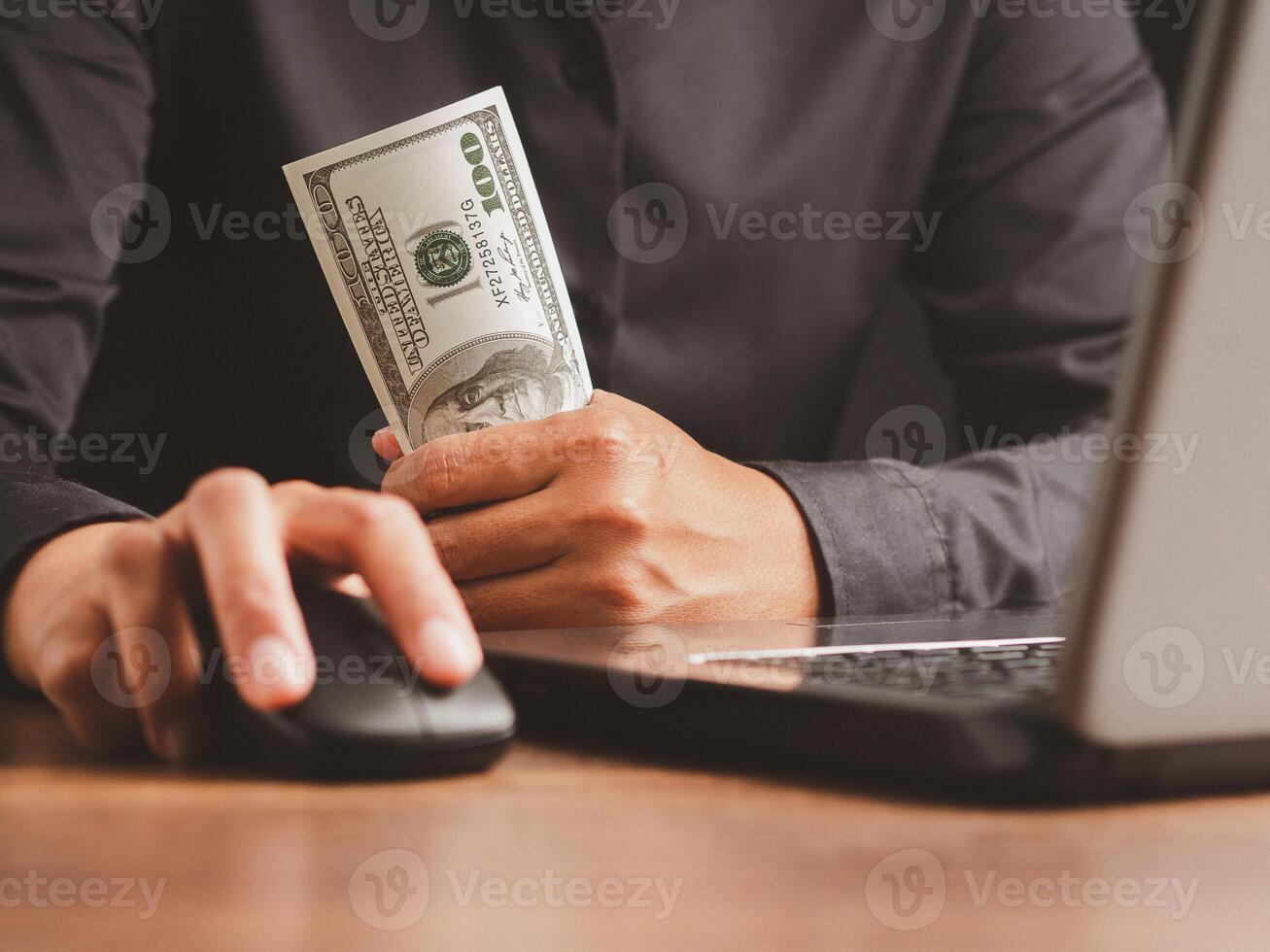 Hand of a businessman in a black shirt holding US dollars banknote and working at the laptop while sitting in the office. photo
