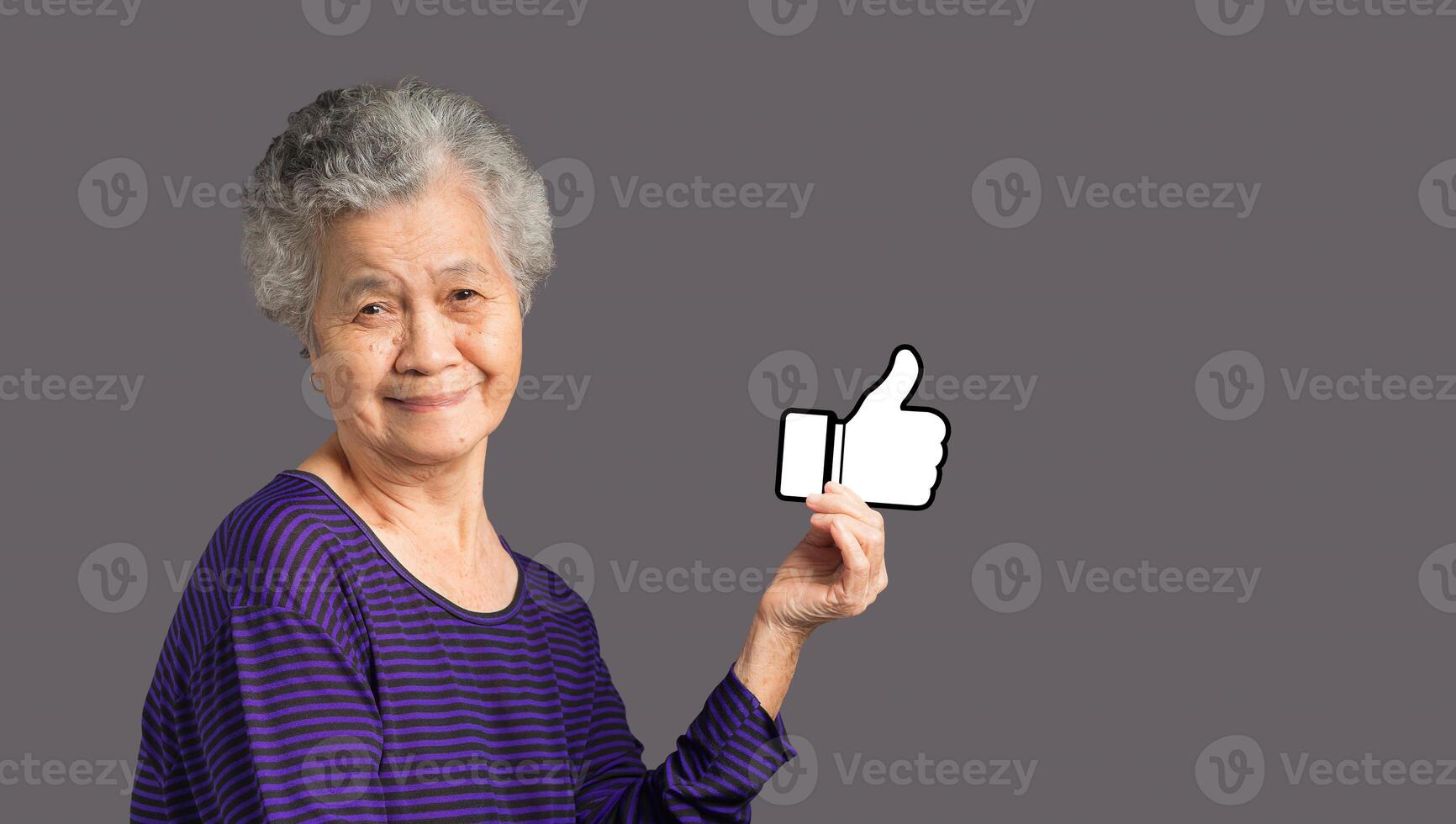 An elderly Asian woman holding a paper thumbs up and looking at the camera with a smile while standing on a gray background photo