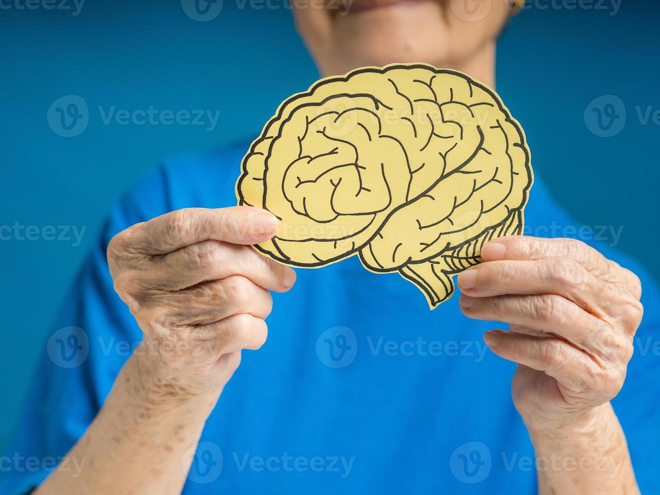 Hands of a senior woman holding a brain shape made from yellow paper against a blue background photo