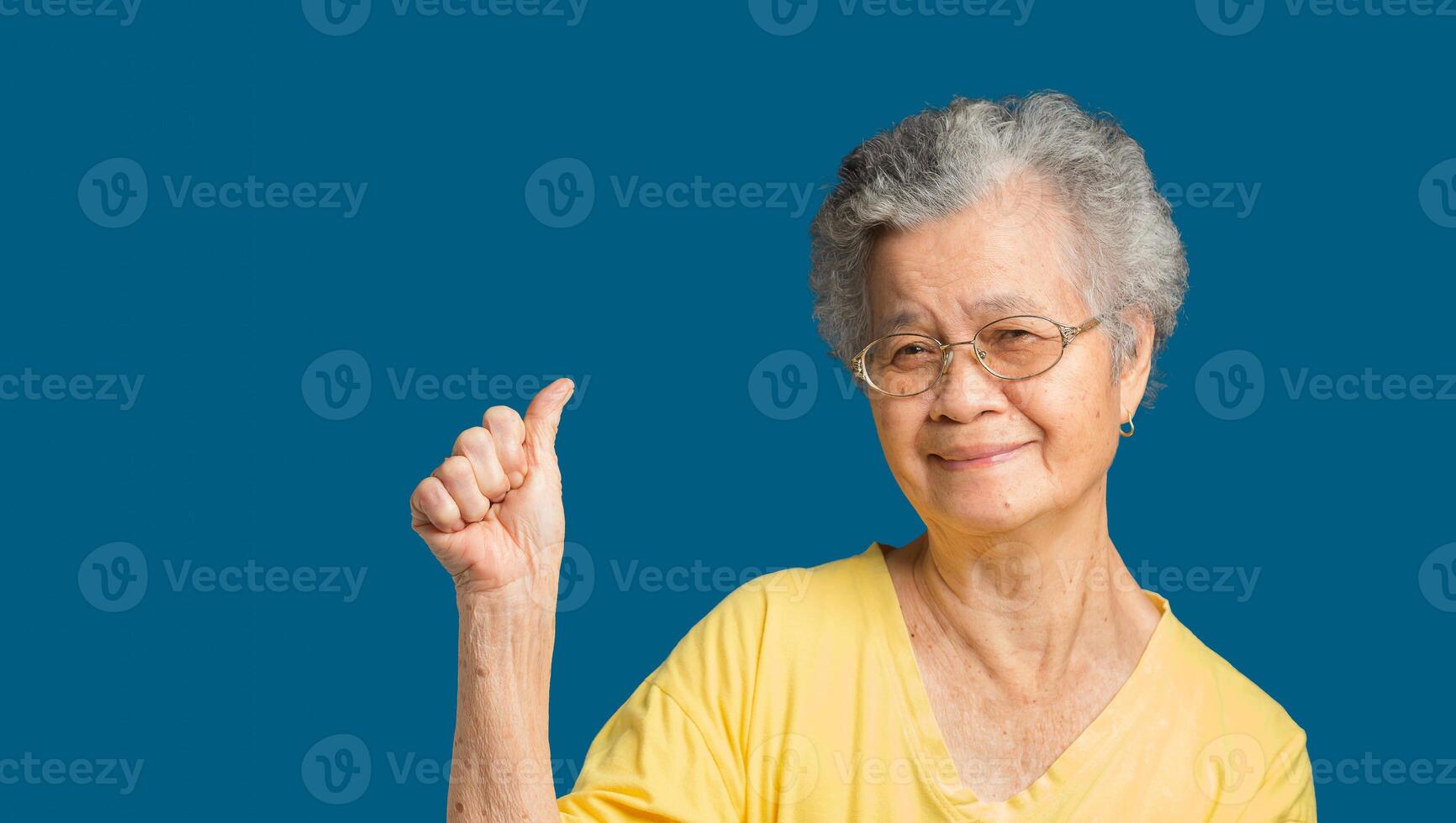 Portrait of an elderly Asian woman showing a thumbs up a symbol and looking at the camera with a smile while standing on a blue background. photo