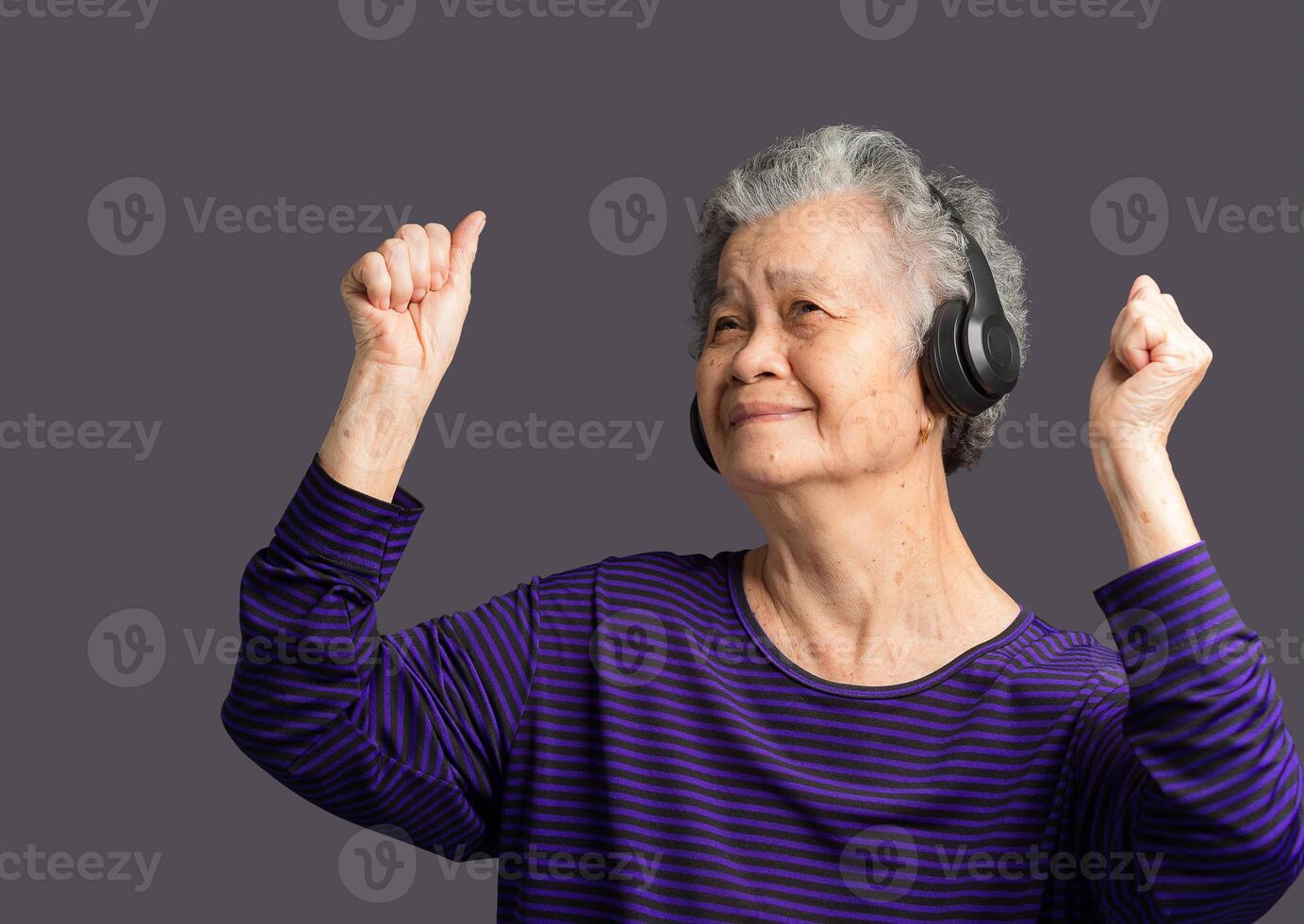 Cheerful elderly Asian woman wireless headphones to listen to a favorite song with a smile while standing on a gray background. photo