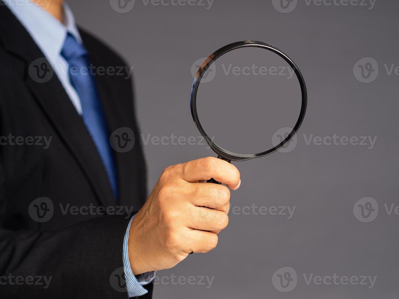 A businessman in a suit holding a magnifying glass while standing over a gray background photo