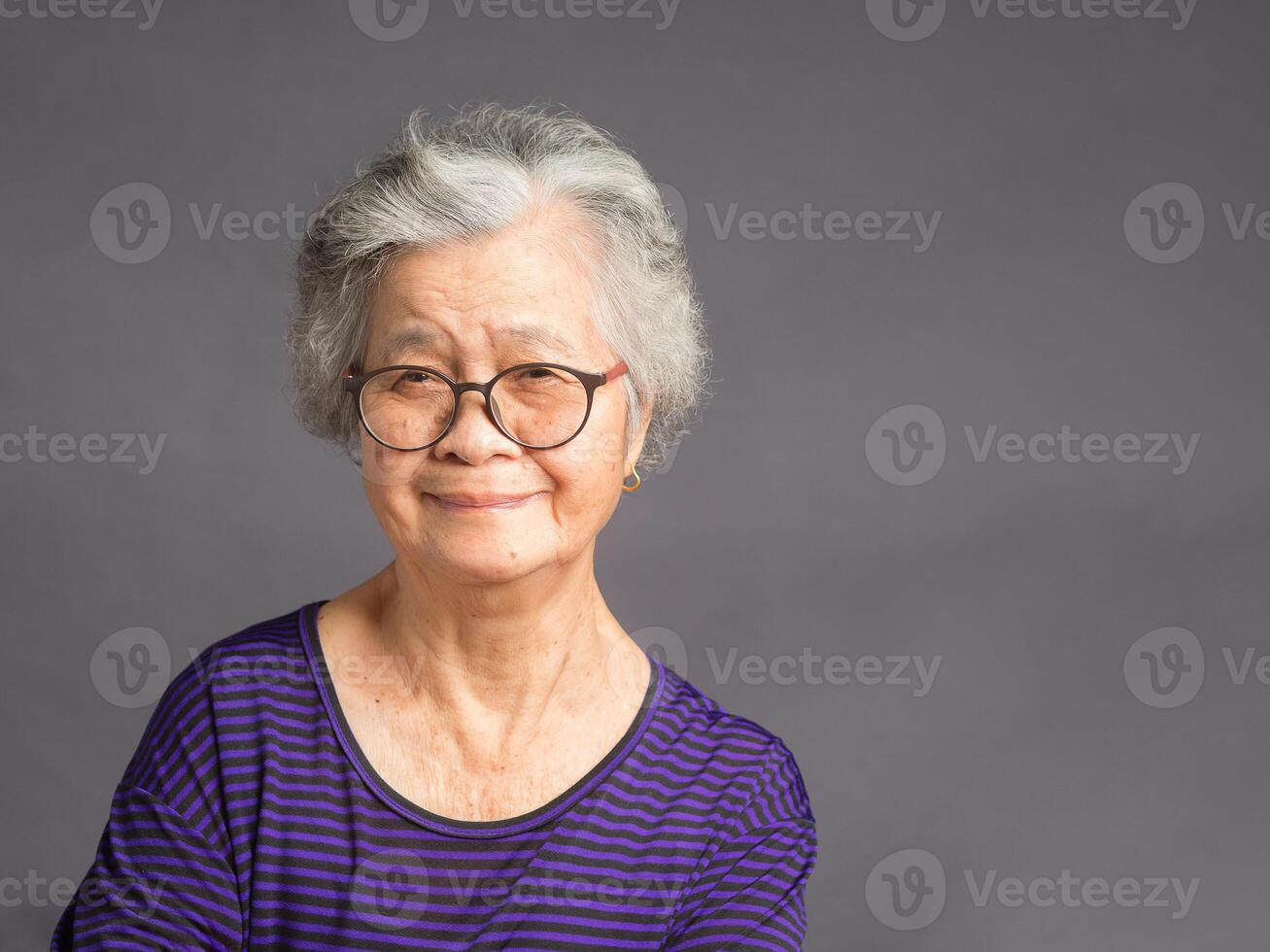 An elderly Asian woman looking at the camera with a smile while standing over a gray background photo